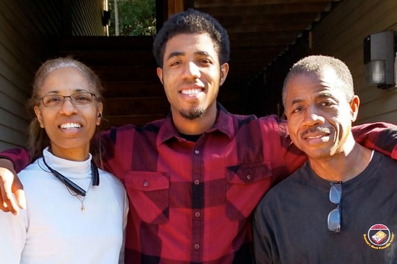 Son standing with his arms over the shoulders of his parents posing for a group shot.