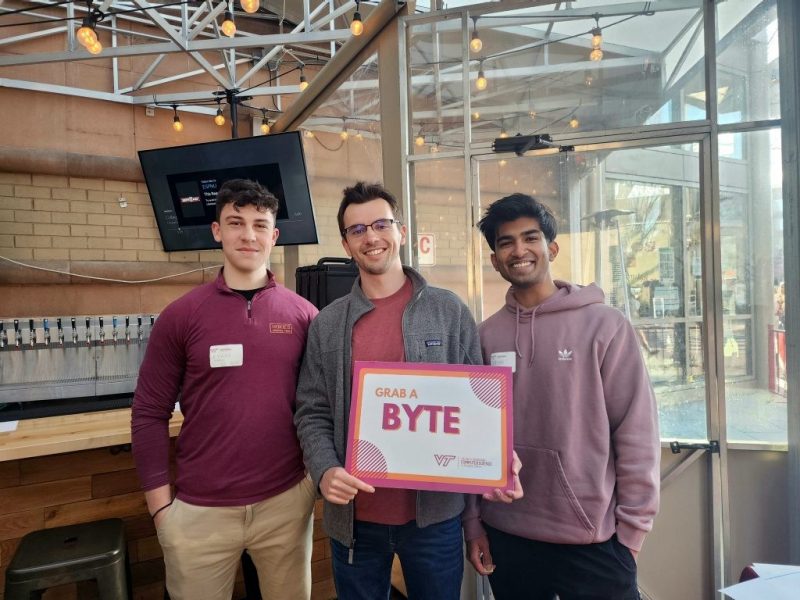 Three alumni smile and hold a sign that says "Grab a Byte"