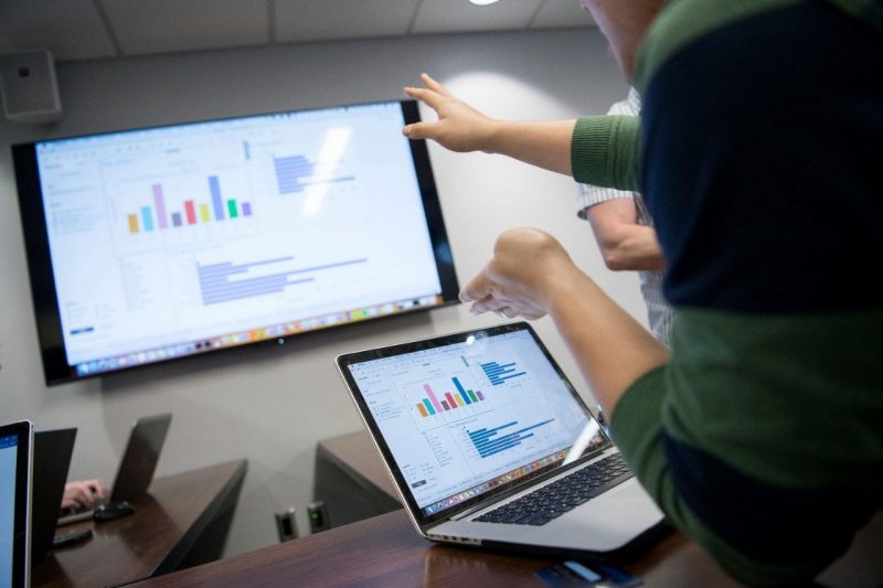 A bar chart on a laptop screen is being displayed on a wall mounted LCD screen.