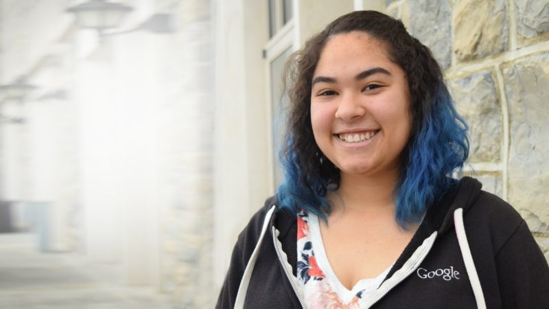 T’Keyaha Sawyers, a 2015 computer science alumna poses for a photos with Hokie stone in the background. She has blue hair and is wearing a Google hoodie.