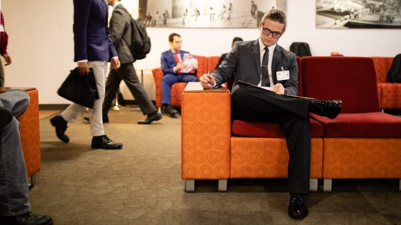 A student sitting on a couch at the SEC Expo with other students walking behind him.