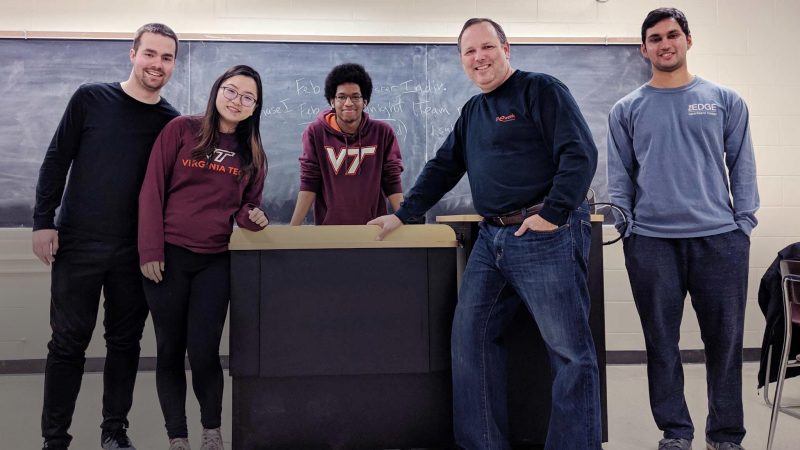 A group of students and a professor pose for a group shot in a classroom.