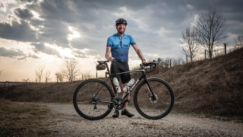 Aaron Goldstein standing with his bike on a gravel path with a dramatic sky behind.