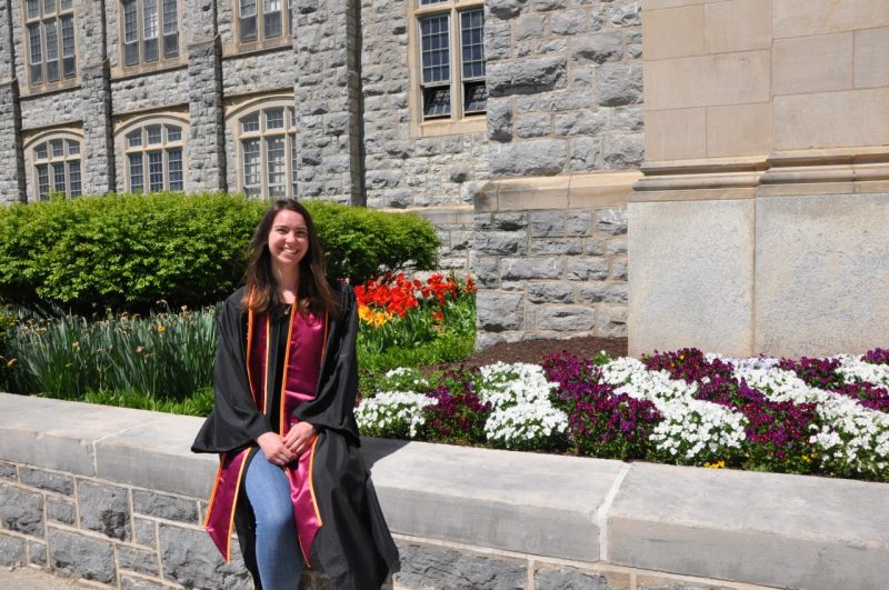 Student with long dark hair, wearing graduation regalia, seated in front of multi color flowers
