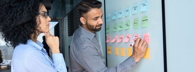 Two multiracial colleagues standing with sticky notes near whiteboard.